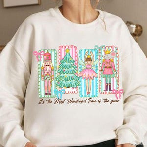 May include: Cream-colored sweatshirt featuring a colorful graphic of a nutcracker, Christmas tree, fairy, and mouse in pink, green, and blue frames. The text "It's the Most Wonderful Time of the year" is printed below the graphic.