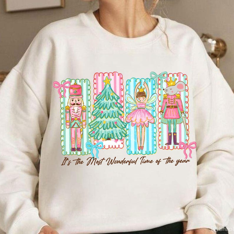May include: Cream-colored sweatshirt featuring a colorful graphic of a nutcracker, Christmas tree, fairy, and mouse in pink, green, and blue frames. The text "It's the Most Wonderful Time of the year" is printed below the graphic.