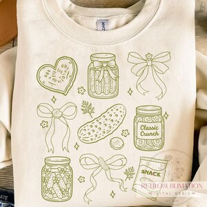May include: Cream-colored sweatshirt featuring a green line-art design of pickles, jars, bows, and a heart-shaped cookie. The design includes the text "Classic Crunch" on a jar and "SNACK" on a bag. The sweatshirt has a crew neck and long sleeves.