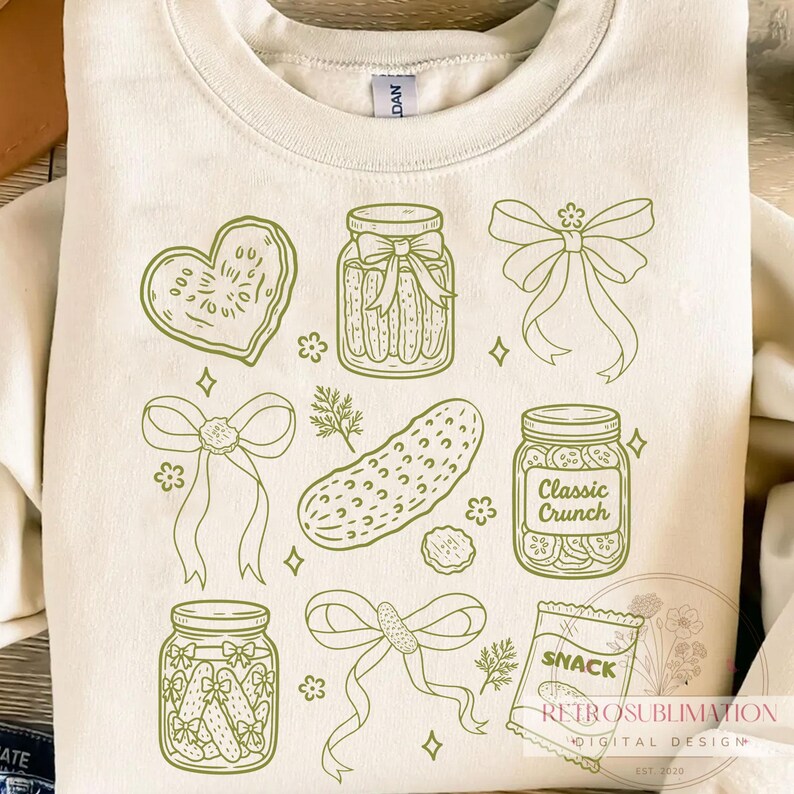 May include: Cream-colored sweatshirt featuring a green line-art design of pickles, jars, bows, and a heart-shaped cookie. The design includes the text "Classic Crunch" on a jar and "SNACK" on a bag. The sweatshirt has a crew neck and long sleeves.