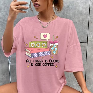 Puede incluir: Camiseta rosa de gran tama&ntilde;o con un gr&aacute;fico de una pila de libros, una taza de caf&eacute; helado y un bocadillo de di&aacute;logo con un coraz&oacute;n. El texto de la camiseta dice "All I need is books & iced coffee."