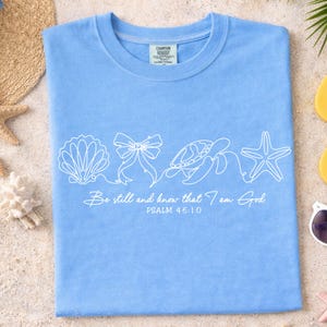 May include: Light blue t-shirt with a white seashell, bow, turtle, and starfish design. The text "Be still and know that I am God" and "Psalm 46:10" are printed below the design. The shirt is laid flat on a sandy surface.