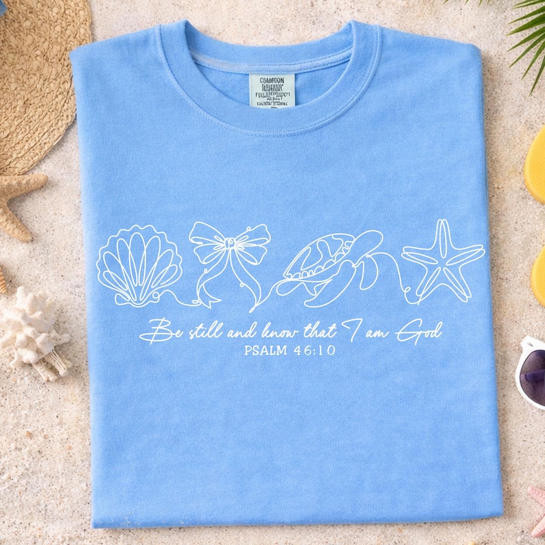 May include: Light blue t-shirt with a white seashell, bow, turtle, and starfish design. The text "Be still and know that I am God" and "Psalm 46:10" are printed below the design. The shirt is laid flat on a sandy surface.