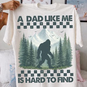 Pode incluir: Camiseta branca com estampa de Bigfoot e o texto "A DAD LIKE ME IS HARD TO FIND". O design apresenta uma silhueta de Bigfoot caminhando em uma floresta com montanhas ao fundo. O texto é em negrito e escuro.