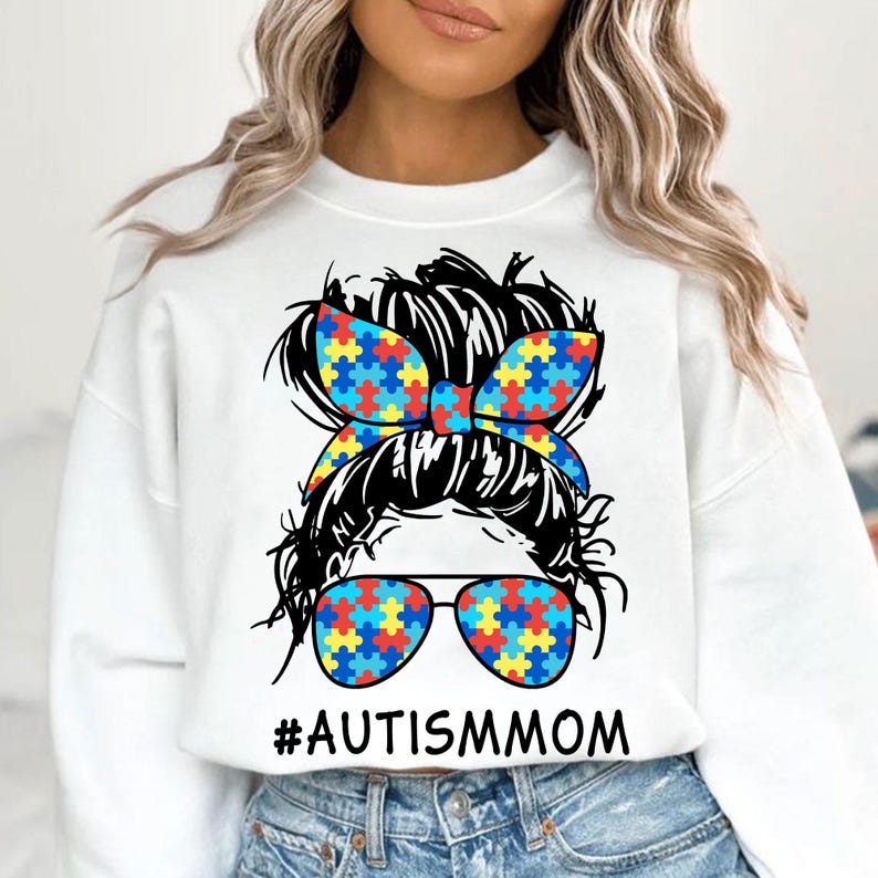 Pu&ograve; includere: Felpa bianca con un'illustrazione in bianco e nero di una donna con uno chignon disordinato. Lo chignon &egrave; decorato con un nastro a motivi di pezzi di puzzle blu, giallo e rosso. La donna indossa occhiali da sole con lo stesso motivo di pezzi di puzzle. Il testo "#AUTISMMOM" &egrave; stampato sotto l'illustrazione.