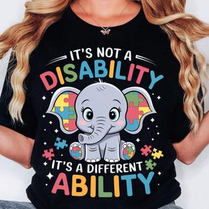 Könnte beinhalten: Schwarzes T-Shirt mit einer bunten Grafik eines Elefanten mit Puzzle-Stücken als Ohren. Der Text "Es ist keine Behinderung, es ist eine andere Fähigkeit" ist in Regenbogenfarben geschrieben.