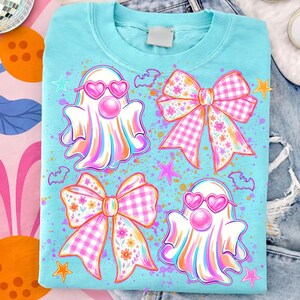 May include: A light blue t-shirt featuring a graphic print of two white ghosts wearing pink heart-shaped sunglasses and blowing pink bubblegum bubbles, accompanied by two large pink and white checkered bows with floral patterns and small stars.