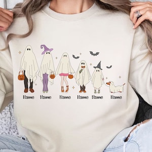 May include: A white sweatshirt with a graphic of a family of ghosts dressed in Halloween costumes. The ghosts are holding pumpkins and a dog is walking next to them. The text "Name" is written below each ghost.