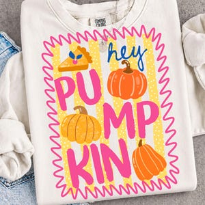 May include: A cream-colored crewneck sweatshirt featuring a graphic with a yellow and white striped background enclosed in a pink wavy border. The design includes three orange pumpkins, a slice of pumpkin pie, and the text "hey PUMPKIN".
