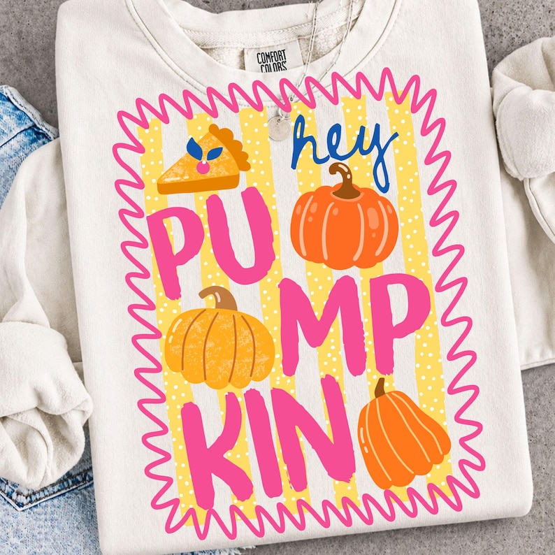 May include: A cream-colored crewneck sweatshirt featuring a graphic with a yellow and white striped background enclosed in a pink wavy border. The design includes three orange pumpkins, a slice of pumpkin pie, and the text "hey PUMPKIN".