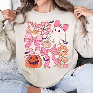 May include: Beige sweatshirt featuring a Halloween-themed design. The design includes pink bows, flowers, a ghost, pumpkins, bats, and a jack-o'-lantern. The graphic is in shades of pink, orange, and black.