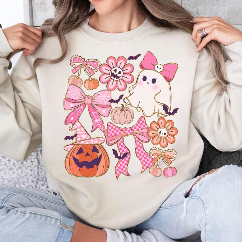 May include: Beige sweatshirt featuring a Halloween-themed design. The design includes pink bows, flowers, a ghost, pumpkins, bats, and a jack-o'-lantern. The graphic is in shades of pink, orange, and black.