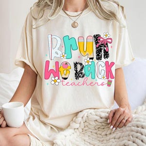 May include: Cream-colored t-shirt with the text "BRU WE BACK teachers" in colorful, playful lettering. The design includes pencils, flowers, and a coffee cup. The shirt is a casual, comfortable style.