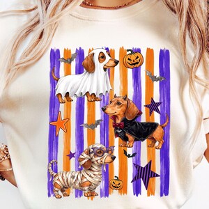 May include: A cream-colored t-shirt featuring a graphic of three dachshunds in Halloween costumes. One dog wears a ghost sheet, one a vampire cape, and one is wrapped like a mummy. The background has vertical purple and orange stripes with bats, pumpkins, and stars.
