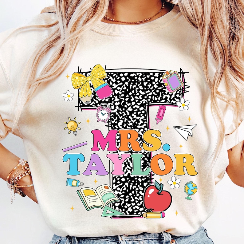 May include: Cream-colored t-shirt featuring the name "MRS. TAYLOR" in colorful letters, with school-themed graphics. The design includes a large letter "T" filled with a black and white pattern, along with illustrations of school supplies.