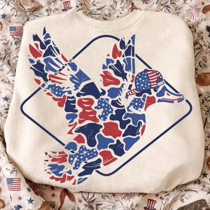 May include: Cream-coloured sweatshirt with a patriotic duck design. The duck is in red, white, and blue camouflage, wearing a baseball cap with an American flag pattern. The design is enclosed within a blue diamond frame.