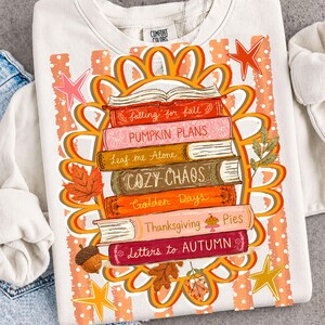 Puede incluir: Una sudadera color crema con un gr&aacute;fico de una pila de libros rodeada por un borde naranja en forma de flor. Los lomos de los libros dicen: falling for fall, PUMPKIN PLANS, Leaf me Alone, COZY CHAOS, Golden Days, Thanksgiving Pies y Letters to AUTUMN.