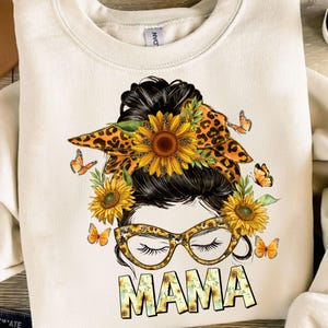 May include: A white sweatshirt with a graphic design of a woman with a messy bun, wearing glasses and a leopard print bandana with sunflowers. The text "MAMA" is written below the graphic in yellow letters with sunflowers.