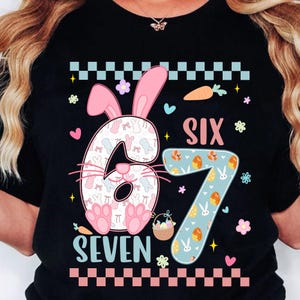 Pode incluir: T-shirt preta com um design colorido com tema de Páscoa. O design apresenta os números "6" e "7" com orelhas de coelho, ovos de Páscoa e as palavras "SIX" e "SEVEN". Elementos decorativos incluem cenouras, flores e uma cesta.