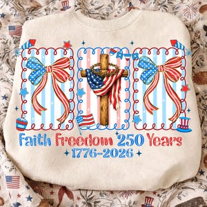 May include: A cream-colored sweatshirt featuring a graphic print with a wooden cross draped in an American flag and a crown of thorns, flanked by two large patriotic bows. The text reads Faith Freedom 250 Years 1776-2026 with firework accents.