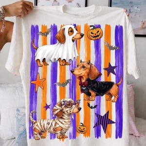 May include: A white t-shirt featuring three dachshunds in Halloween costumes: one as a ghost, one as a vampire with a cape, and one as a mummy. The background has vertical purple and orange brushstrokes, orange and purple stars, bats, and pumpkins.