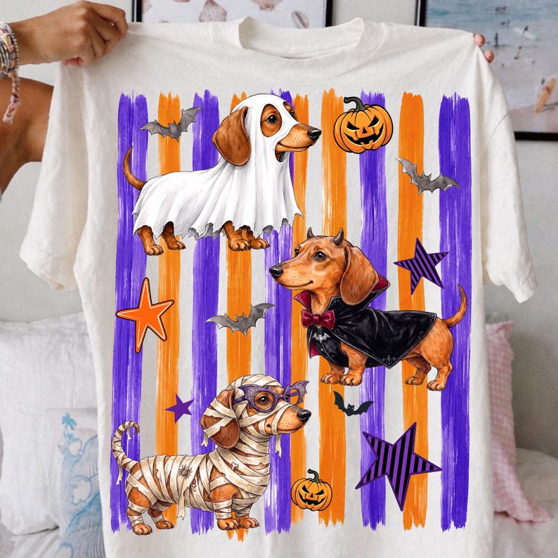 May include: A white t-shirt featuring three dachshunds in Halloween costumes: one as a ghost, one as a vampire with a cape, and one as a mummy. The background has vertical purple and orange brushstrokes, orange and purple stars, bats, and pumpkins.