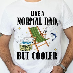 Puede incluir: Camiseta blanca con un diseño gráfico negro que dice "Como un padre normal, pero más guay". El gráfico también incluye una imagen de dibujos animados de una silla de playa, una nevera portátil y un pez.