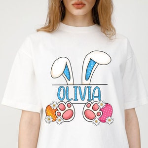 May include: White t-shirt with a bunny ear design and the name "Olivia" printed on it. The bunny ears have blue and white polka dots. The bunny feet are pink with white paw prints. The bunny feet are surrounded by orange and pink Easter eggs with white polka dots and white daisies.