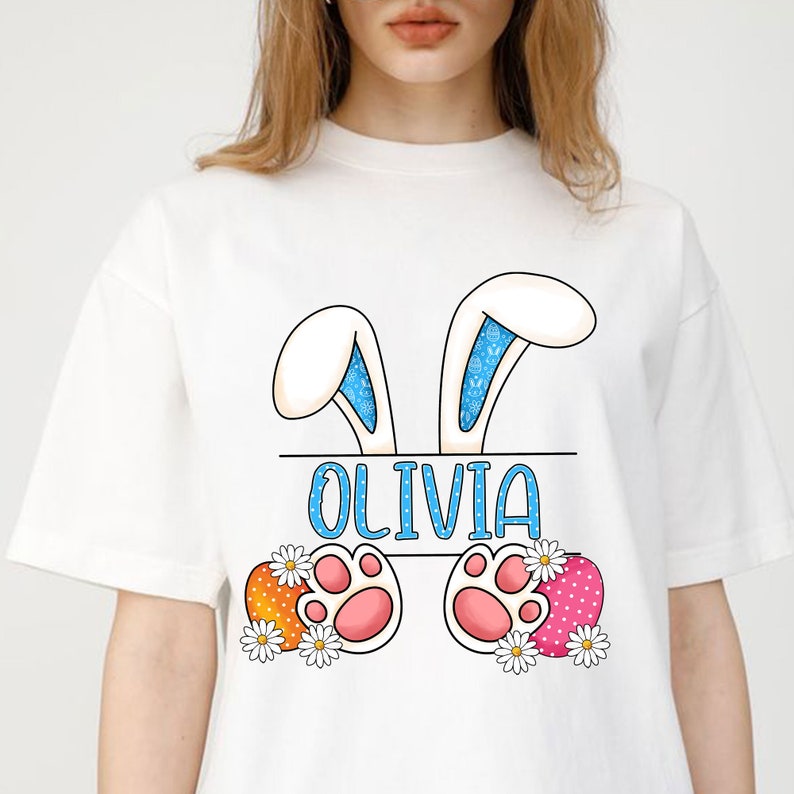 May include: White t-shirt with a bunny ear design and the name "Olivia" printed on it. The bunny ears have blue and white polka dots. The bunny feet are pink with white paw prints. The bunny feet are surrounded by orange and pink Easter eggs with white polka dots and white daisies.