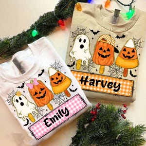 May include: Two Halloween-themed sweatshirts. One white sweatshirt features a ghost, pumpkin, and candy corn popsicle design with the name "Emily". The other is beige and features similar designs with the name "Harvey". Both have spiderwebs and bats.