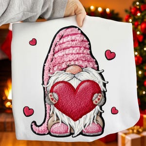 Könnte beinhalten: Weißes Kissen mit einem gestickten Zwerg, der ein rotes Herz hält. Der Zwerg hat einen rosa Hut und einen langen weißen Bart. Kleine rote Herzen sind um den Zwerg herum verstreut. Das Kissen wird von einer Person gehalten.