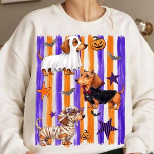 May include: A cream-colored crewneck sweatshirt featuring a graphic print of three dachshunds in Halloween costumes. One dog wears a white ghost sheet, one wears a black vampire cape with a red bow tie, and one is wrapped in white bandages like a mummy.