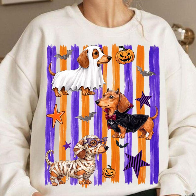 May include: A cream-colored crewneck sweatshirt featuring a graphic print of three dachshunds in Halloween costumes. One dog wears a white ghost sheet, one wears a black vampire cape with a red bow tie, and one is wrapped in white bandages like a mummy.