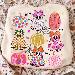 May include: A cream-colored sweatshirt featuring a colorful graphic print of four ghosts wearing bows and four pumpkins. The ghosts have patterns including leopard print, checkerboard, floral, and daisies. The pumpkins feature polka dots, stripes, and hearts.