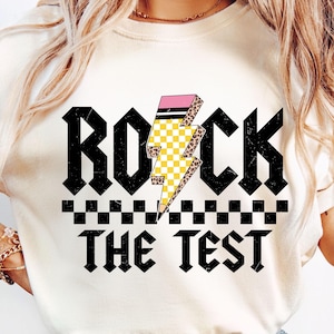 May include: White t-shirt with a black and white checkered pattern and the text "Rock the Test" in black. A yellow and black lightning bolt with pink and leopard print accents is behind the text.