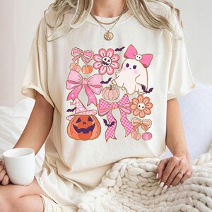 May include: Cream-colored t-shirt featuring a Halloween-themed graphic. The design includes pink bows, flowers, pumpkins, a ghost, and bats. The pumpkin wears a witch hat. The overall design has a cute, vintage aesthetic.
