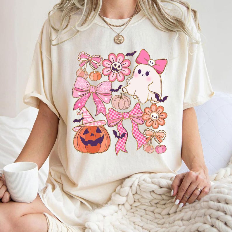 May include: Cream-colored t-shirt featuring a Halloween-themed graphic. The design includes pink bows, flowers, pumpkins, a ghost, and bats. The pumpkin wears a witch hat. The overall design has a cute, vintage aesthetic.