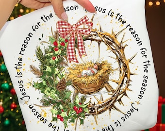 May include: White square pillow with a Christmas theme. The pillow features a baby in a manger, a crown of thorns, and a red and white plaid bow. Text around the image reads "Jesus is the reason for the season."