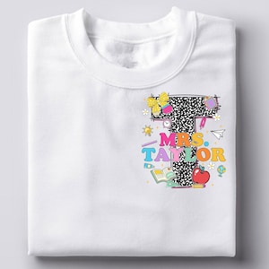 May include: White sweatshirt featuring a colorful graphic design. The design includes the text "MRS. TAYLOR" in vibrant colors, a large letter "T" with a black and white pattern, and various school-themed illustrations like an apple, books, and a globe.