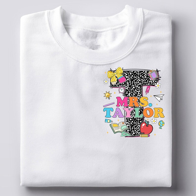 May include: White sweatshirt featuring a colorful graphic design. The design includes the text "MRS. TAYLOR" in vibrant colors, a large letter "T" with a black and white pattern, and various school-themed illustrations like an apple, books, and a globe.