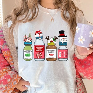 Puede incluir: Camiseta blanca con un dise&ntilde;o navide&ntilde;o con cuatro botellas con tapas y etiquetas festivas. Las etiquetas dicen "Spiced Vanilla Versed", "Peppermint Mocha Propofol", "Hazelnut Haldol" y "Apple Crisp Ativan".