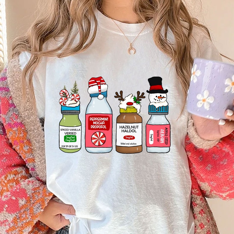 Puede incluir: Camiseta blanca con un dise&ntilde;o navide&ntilde;o con cuatro botellas con tapas y etiquetas festivas. Las etiquetas dicen "Spiced Vanilla Versed", "Peppermint Mocha Propofol", "Hazelnut Haldol" y "Apple Crisp Ativan".