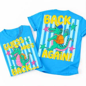 May include: Two bright blue t-shirts featuring a cartoon green crocodile wearing a yellow and blue cap and an orange backpack. The shirts display the text "GUESS WHO'S BACK..." and "BACK AGAIN!" over a light blue striped background with colorful stars.