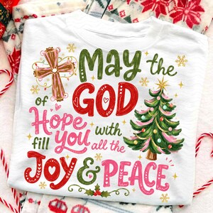May include: A white crew neck t-shirt featuring a graphic print with a brown cross, a green Christmas tree, and the text: May the GOD of Hope fill you with all the JOY & PEACE. The design includes small gold snowflakes and red heart accents.