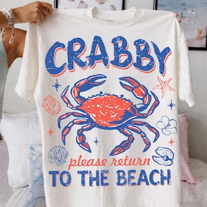 以下が含まれることがあります： クリーム色のTシャツ。青と赤のひび割れたカニのグラフィックと「CRABBY」と「please return TO THE BEACH」の文字。デザインにはヒトデ、貝殻、花が含まれています。Tシャツは人が持っています。