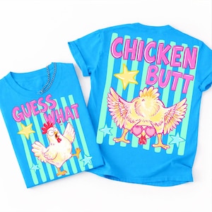 May include: Two blue t-shirts with chicken-themed graphics. One shirt says "GUESS WHAT" with a cartoon chicken. The other says "CHICKEN BUTT" with a chicken wearing heart-shaped sunglasses. Both shirts have star and stripe designs.