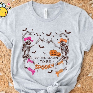 May include: A grey t-shirt featuring a graphic of two skeletons wearing cowboy hats and boots, dancing with pumpkins. The text "Tis' the season to be spooky" is printed on the shirt.