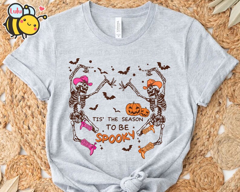 May include: A grey t-shirt featuring a graphic of two skeletons wearing cowboy hats and boots, dancing with pumpkins. The text "Tis' the season to be spooky" is printed on the shirt.