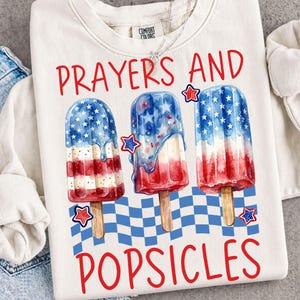 May include: White sweatshirt with the words "Prayers and Popsicles" in red. The design features three popsicle illustrations in red, white, and blue, with star accents, evoking a patriotic theme. A blue and white checkered pattern is at the bottom.