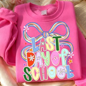 May include: A bright pink sweatshirt with the text "LAST DAY OF SCHOOL" in colorful, playful lettering. The design includes a large rainbow-colored bow and various star and flower accents, creating a cheerful, celebratory vibe.